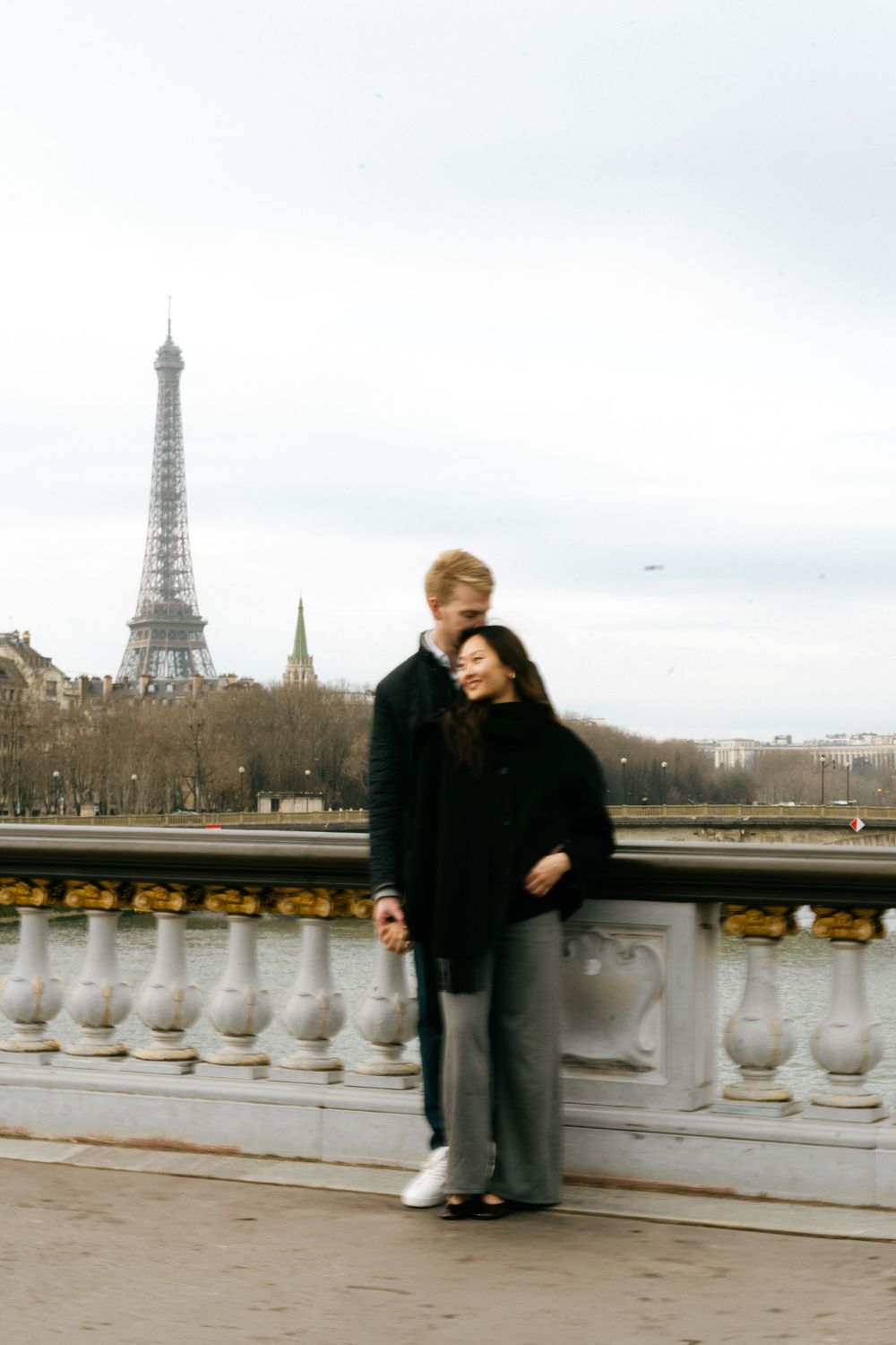 A couple shares a romantic moment on a bridge overlooking the Eiffel Tower in Paris on an overcast day. Pont Alexandre III.