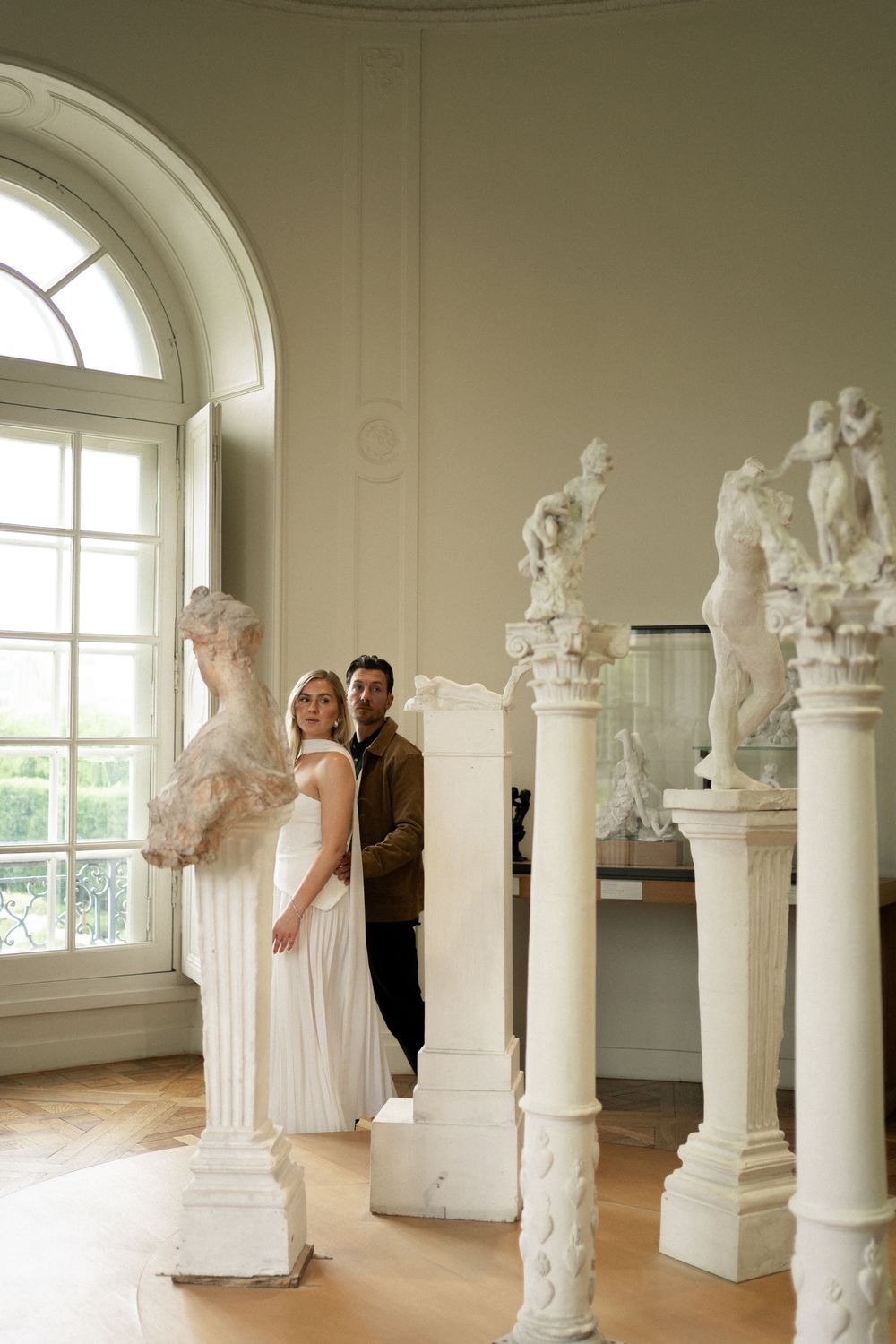 Two people pose elegantly among classical white marble sculptures and columns in a sunlit museum gallery with arched windows.