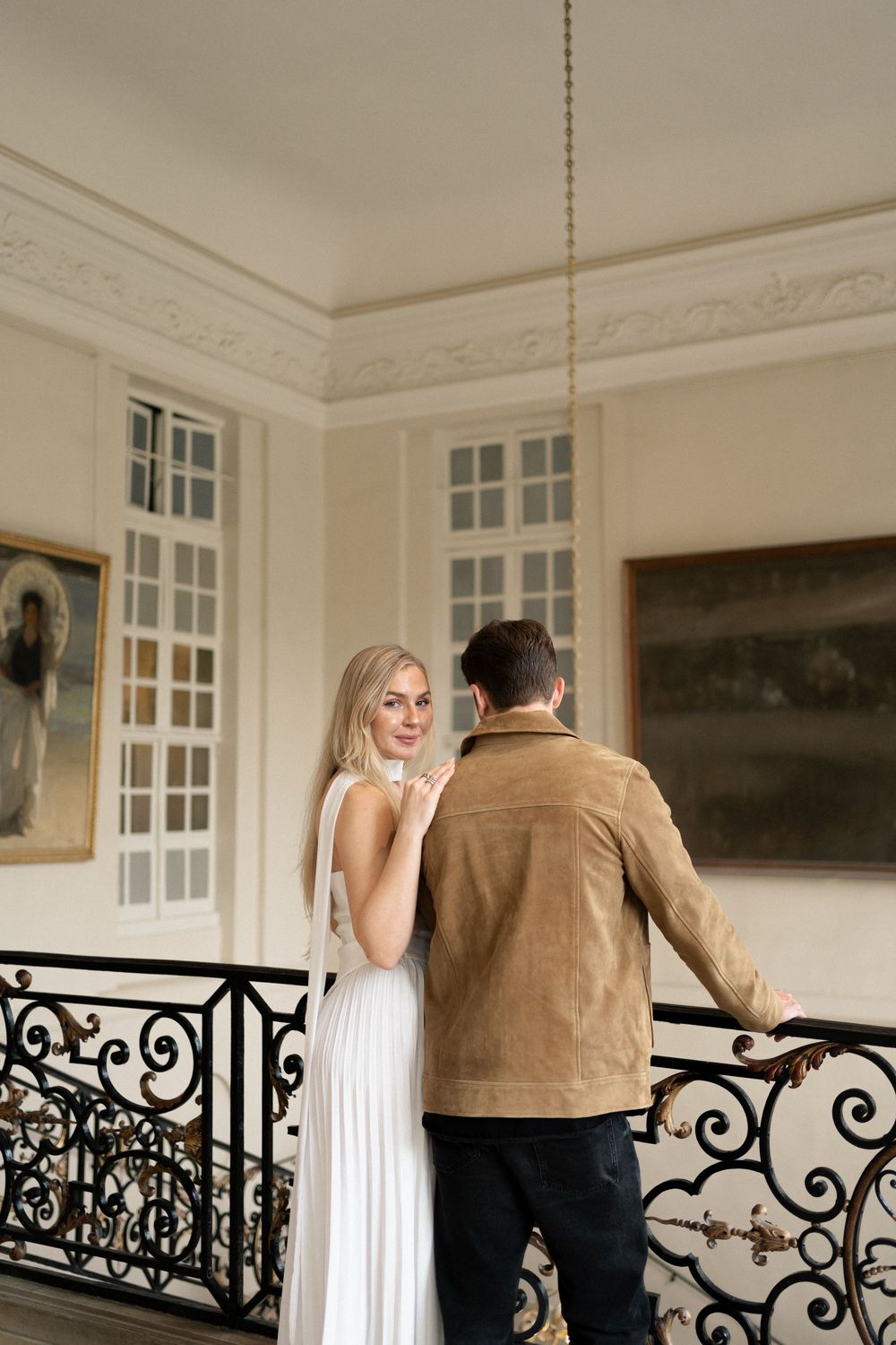 A couple stands on an elegant staircase with ornate iron railings in a grand interior with tall French windows.