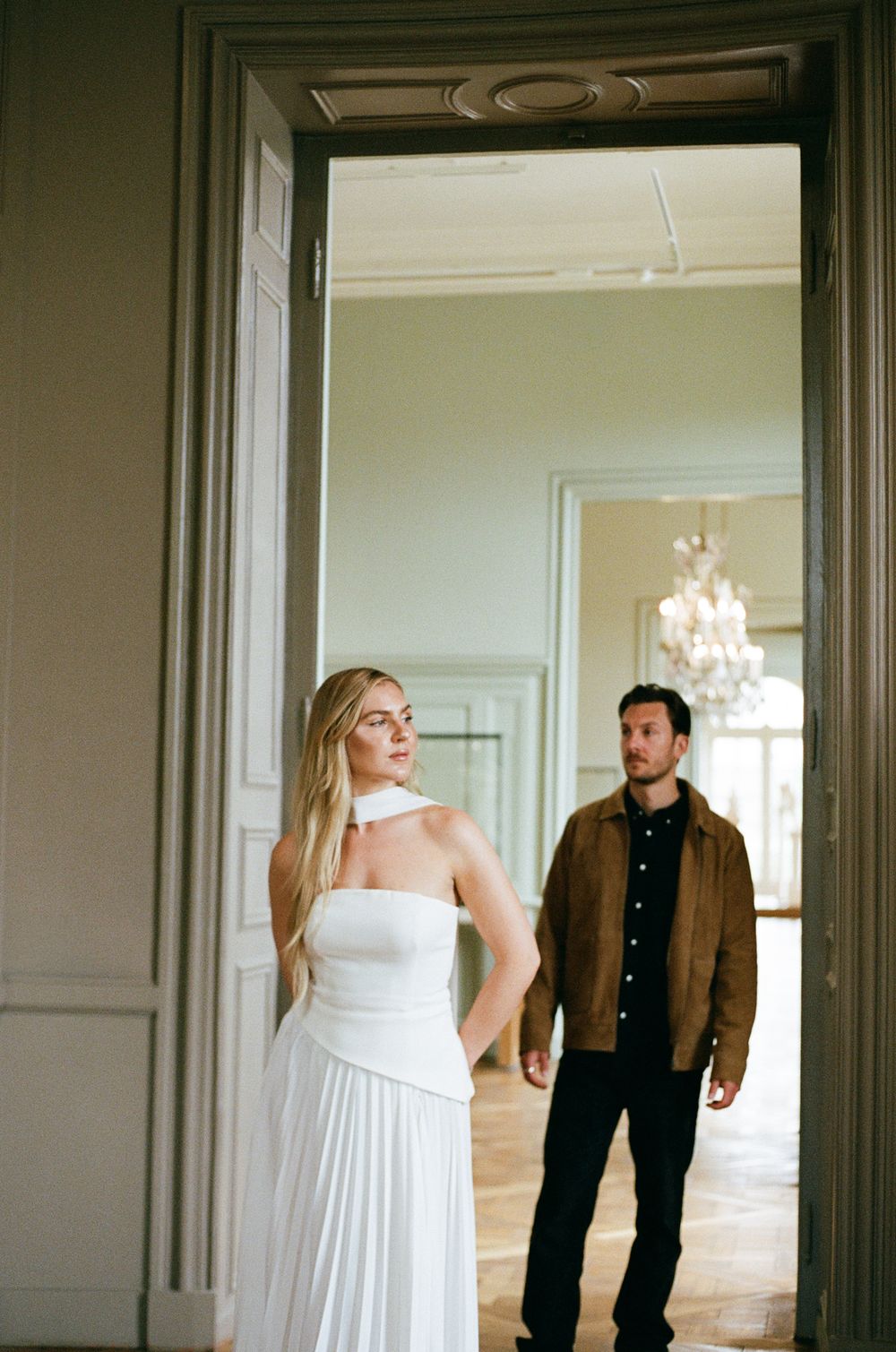 A person in a flowing white gown stands in an elegant room with chandeliers while another person waits in the doorway.