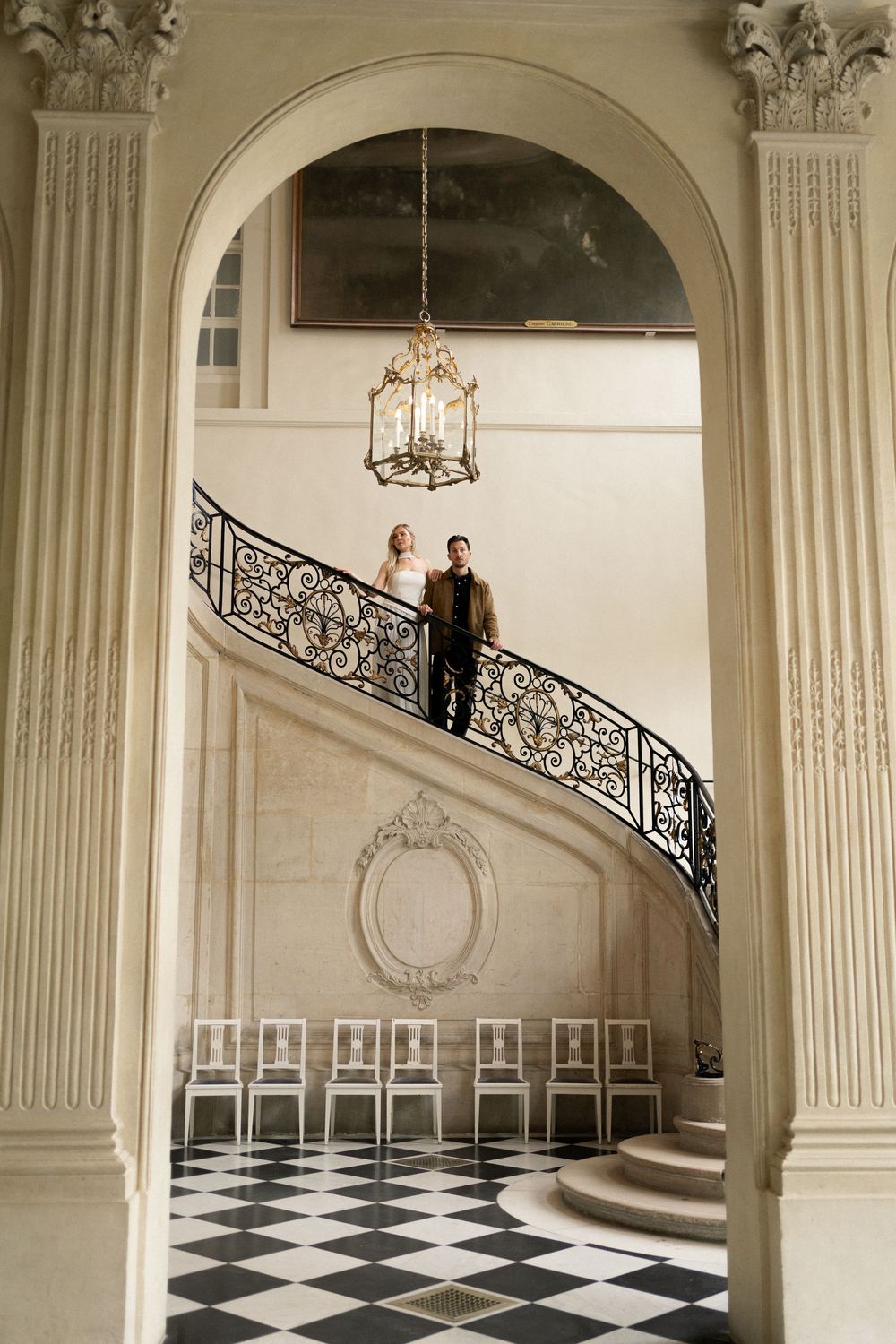 Elegant grand staircase with ornate iron railings and checkerboard marble floors in a classical Baroque palace interior.