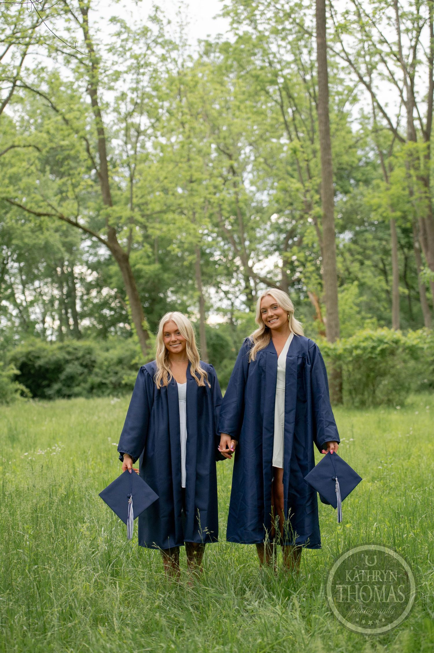 Two graduates in navy blue caps and gowns stand together in a sunny green field with trees in the background.