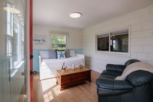 Bright sunlit living room with white walls features a black leather sofa, wooden storage trunk, and large windows.