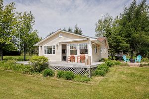 Charming white cottage with a wraparound deck sits on a grassy lawn surrounded by trees in a peaceful countryside setting.