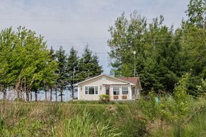 White single-story cottage with bay windows sits atop grassy hill surrounded by evergreen trees and natural vegetation.