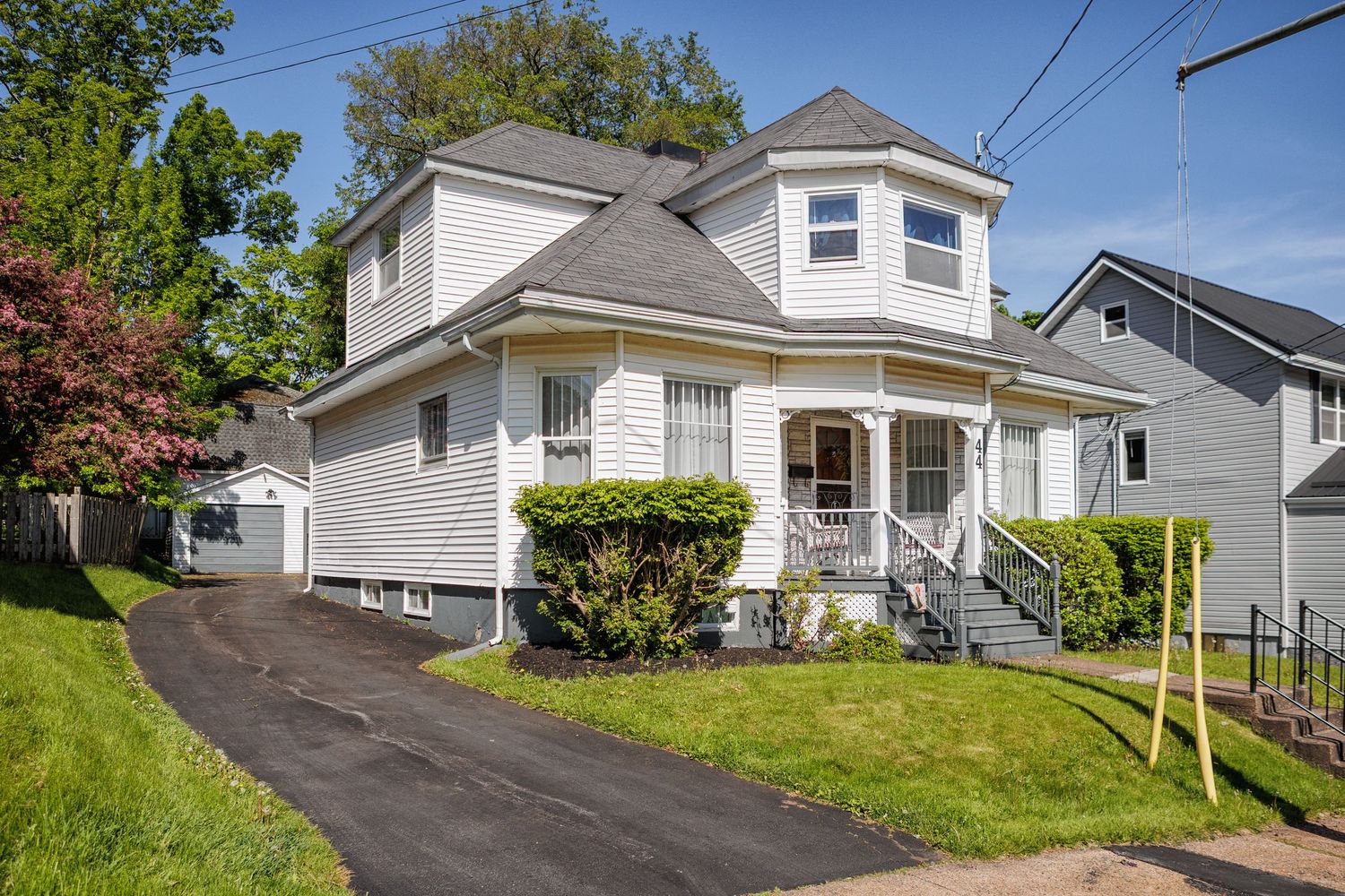 Charming white Victorian-style house with turret and front porch sits on a sloped lawn with black driveway.