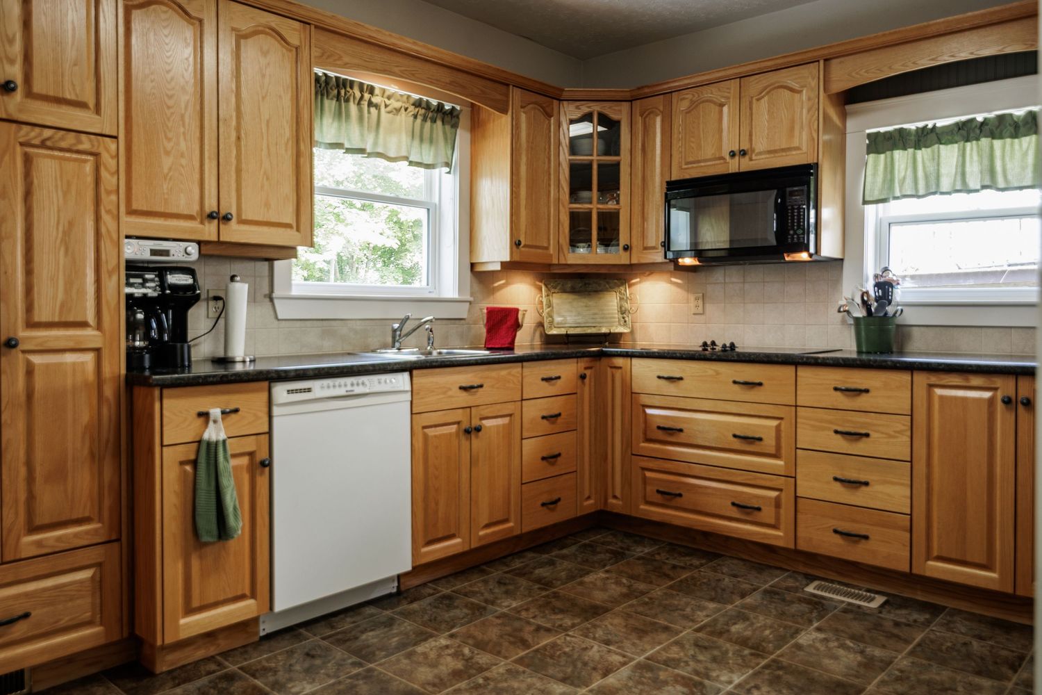 Wraparound honey oak kitchen cabinets surround white appliances with sage green curtains and a beige tiled countertop.