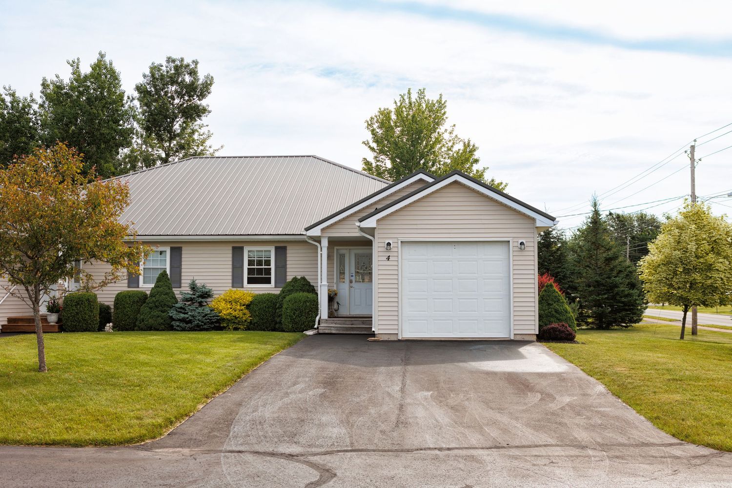 Single-story ranch home with beige siding, white garage, covered porch, and lush green lawn surrounded by mature trees.