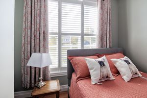 Cozy bedroom with coral bedding, decorative pillows, white plantation shutters, floral curtains, and a wooden bedside table with lamp.