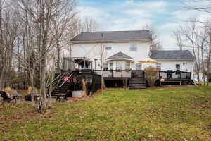 White two-story house with elevated wooden deck and stairs on sloping backyard surrounded by bare winter trees.