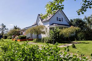 White farmhouse style home nestled on a green grassy hillside with mature trees and shrubs in a sunny rural setting.