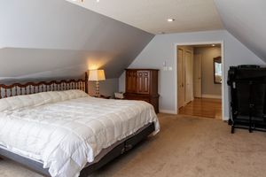 Cozy attic bedroom with sloped ceiling features white bedding, beige carpet, and warm lighting for a welcoming atmosphere.