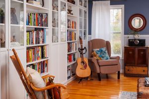 Cozy reading nook with white built-in bookshelves, wooden rocking chair, brown armchair, acoustic guitar and hardwood floors.