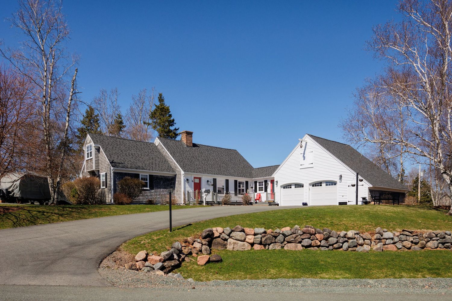 White Cape Cod home with attached garage, stone wall landscaping, and circular driveway under clear blue sky.