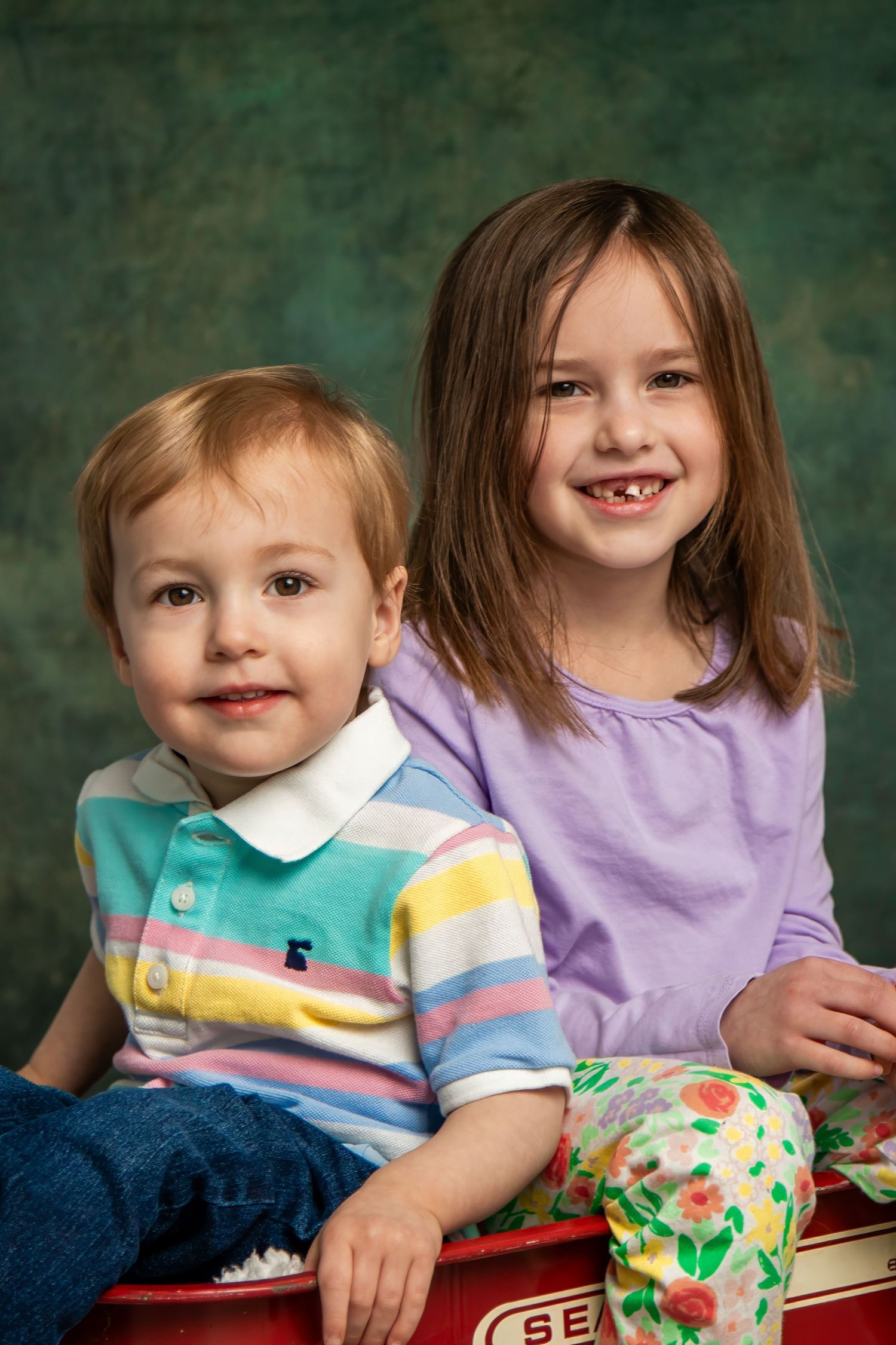 Two children in casual clothing sit together for a studio portrait against a green backdrop.