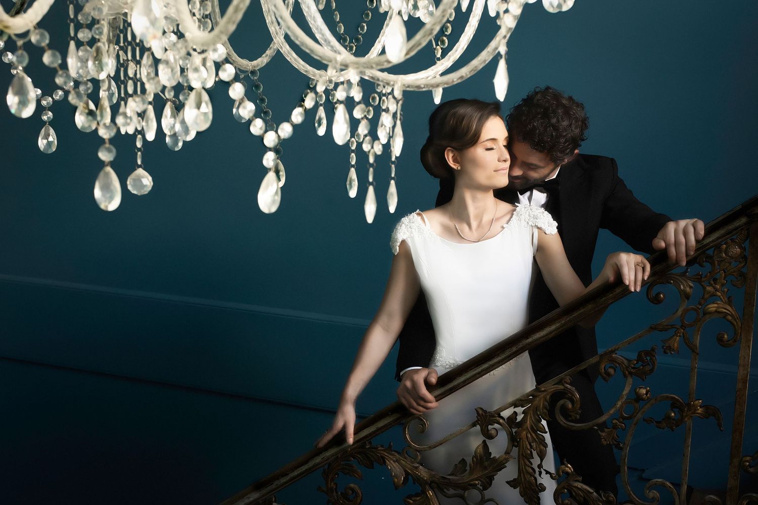Romantic couple poses beneath elegant crystal chandeliers in a moody blue-toned room setting.