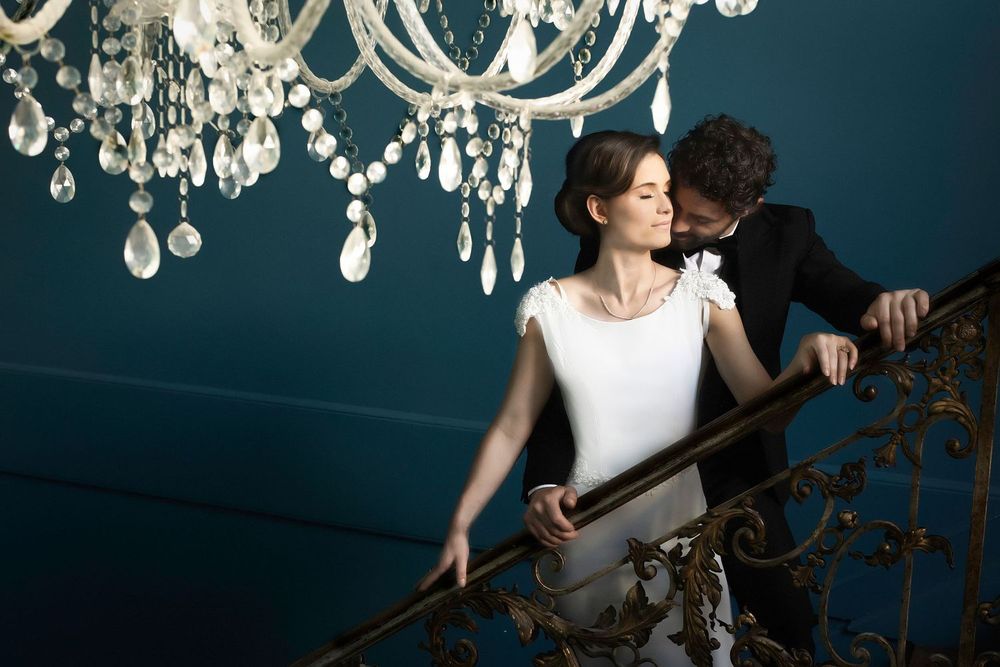 Romantic couple poses beneath elegant crystal chandeliers in a moody blue-toned room setting.