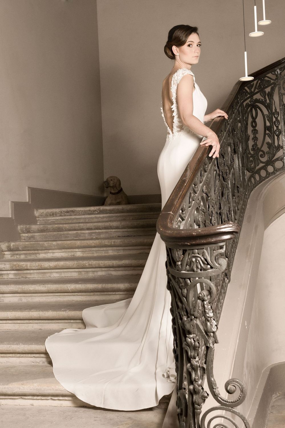 Elegant wedding gown with low back and long train photographed on grand staircase with ornate railing.