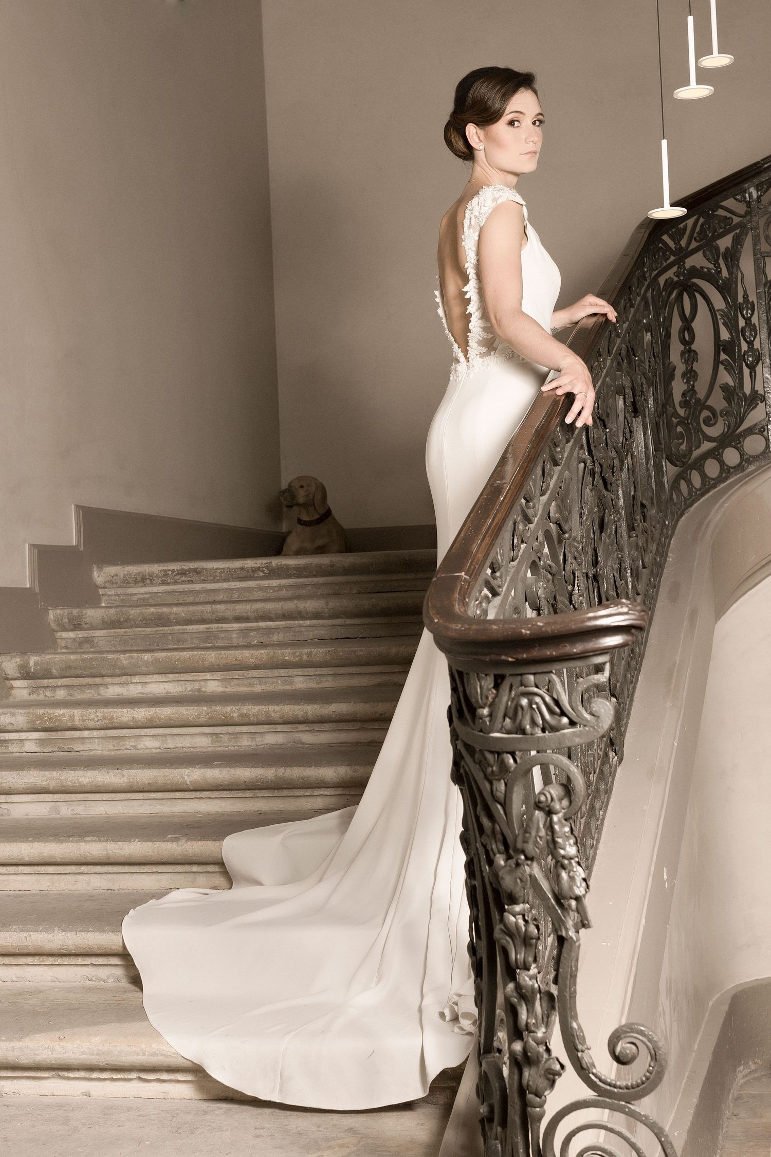Elegant wedding gown with low back and long train photographed on grand staircase with ornate railing.