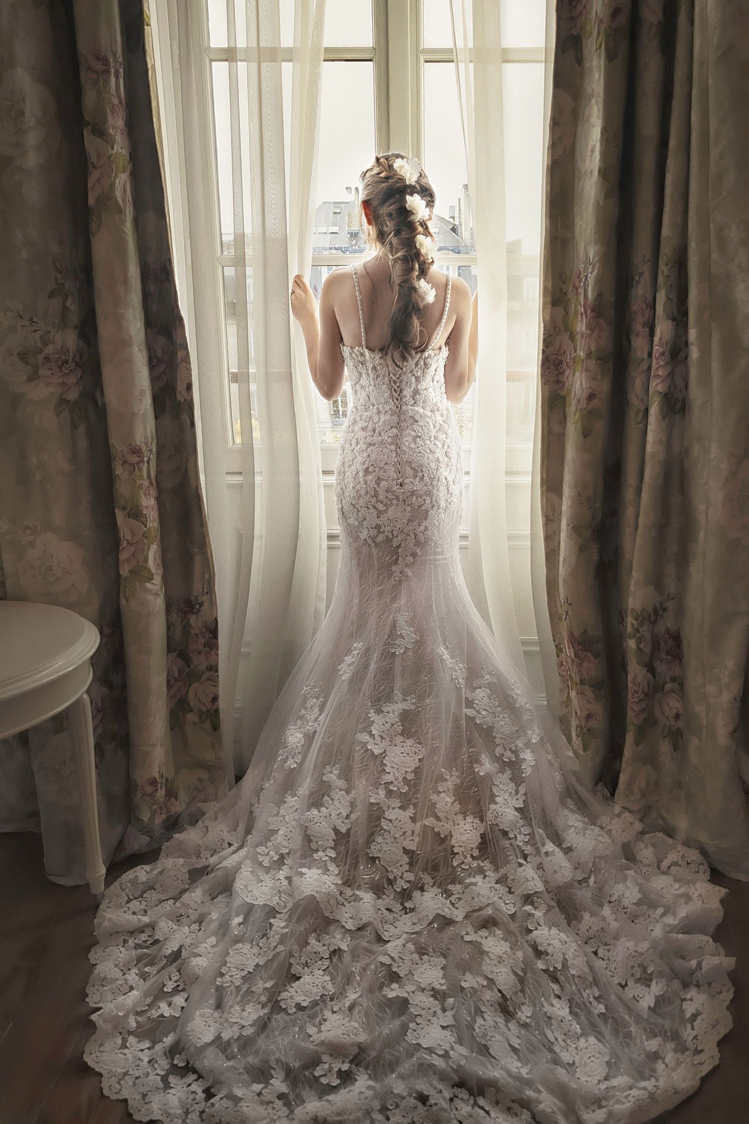 Backlit silhouette of lace wedding gown with dramatic floral train against window and curtains.