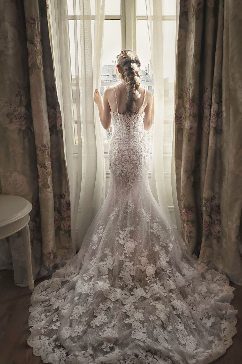 Backlit silhouette of lace wedding gown with dramatic floral train against window and curtains.