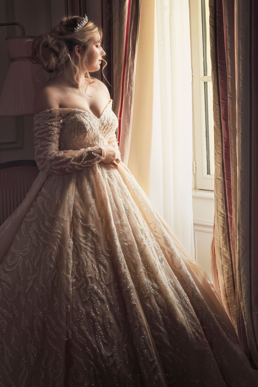 A romantic silhouette by a window shows an off-shoulder vintage wedding gown with intricate lace and a flowing ballgown skirt.