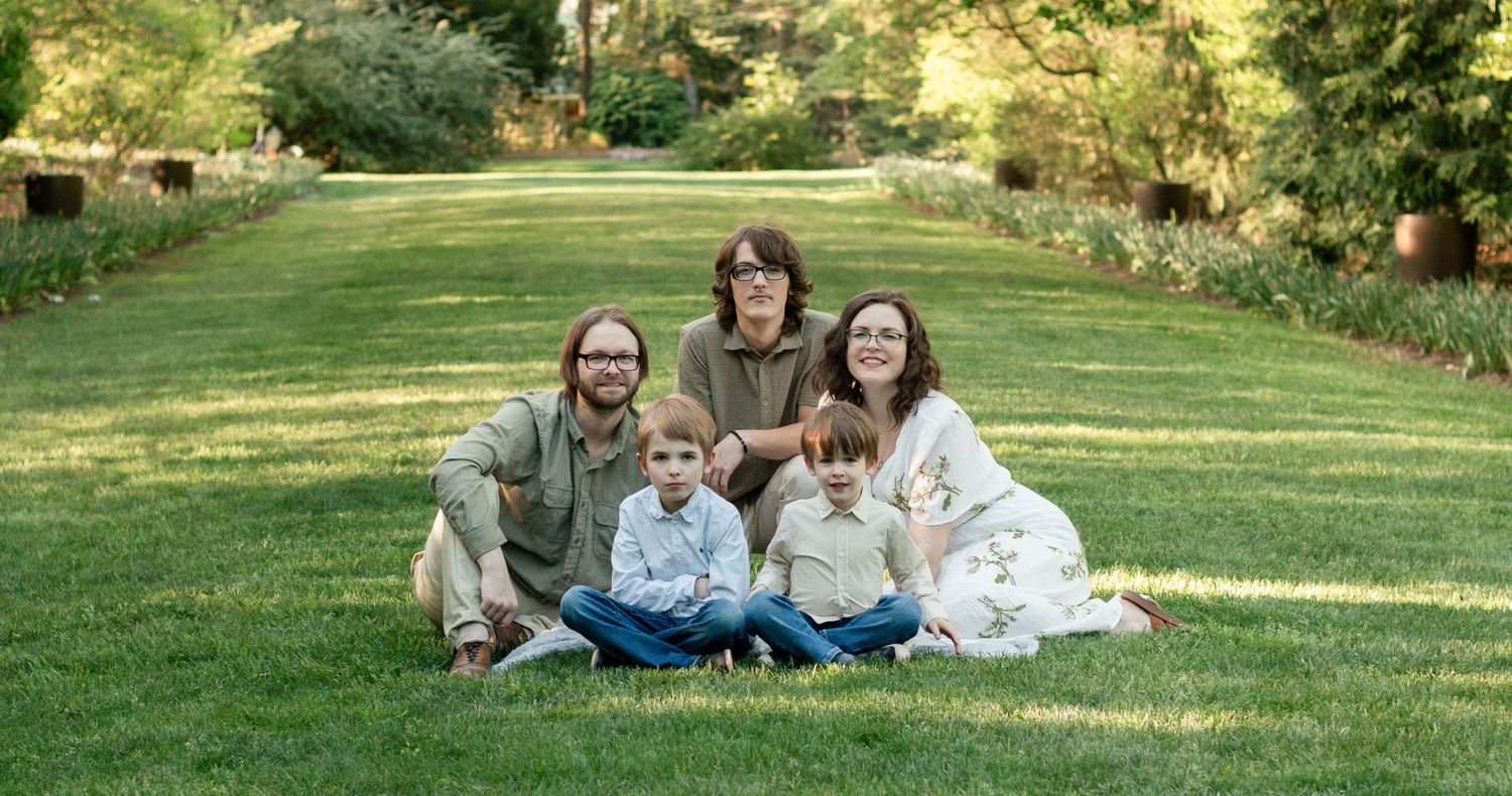 A group sits together on lush green grass in a garden setting with sunlit trees in the background.