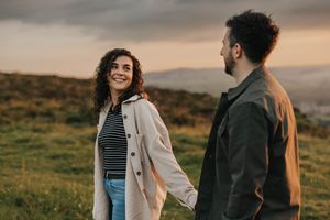 Couple stands together in romantic sunset moment on grassy hillside with golden mountain views in background. By Lincolnshire photographers.