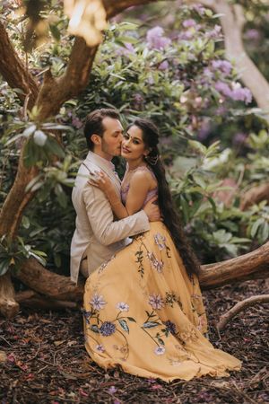 A couple in elegant attire embraces beneath flowering trees, with the woman wearing a yellow floral gown. By Lincolnshire photographers.
