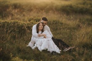 A couple in white clothing embrace while sitting in a sunlit meadow at golden hour among tall grass.