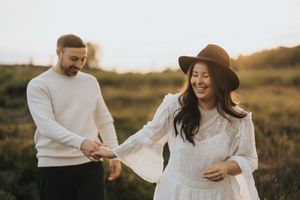 Two people in white outfits embrace and laugh together during a sunset engagement photo shoot in a golden field.