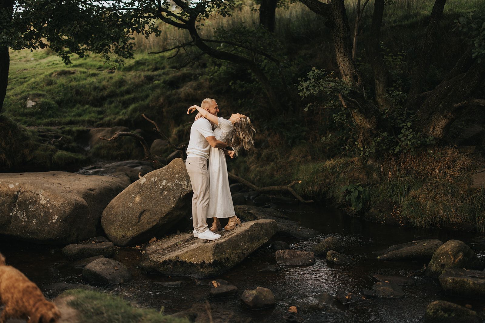 A couple in white outfits embrace while standing on rocks beside a serene stream in a wooded setting. Captured by Lincolnshire photographers