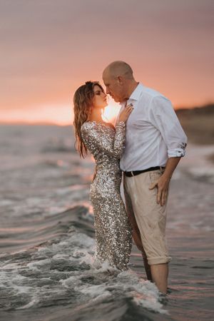 Romantic couple embraces in sparkling sequin dress and casual attire while standing in ocean waves at sunset.