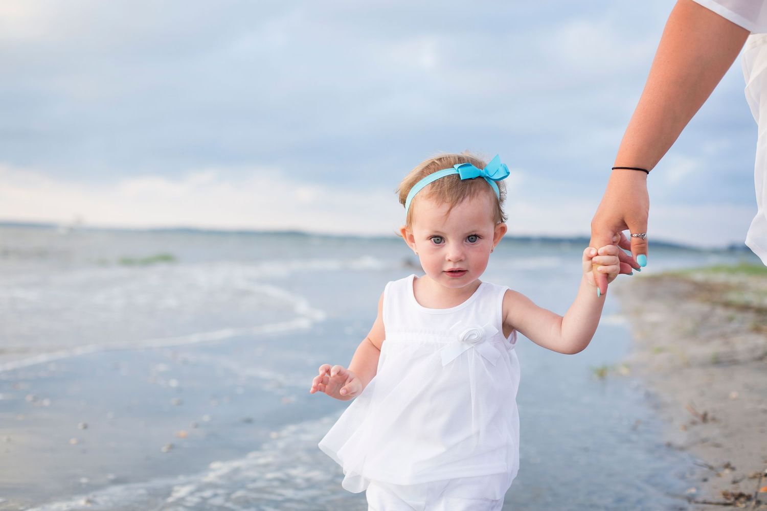 A small child in a white dress walks along the beach while holding an adult's hand at the shoreline.