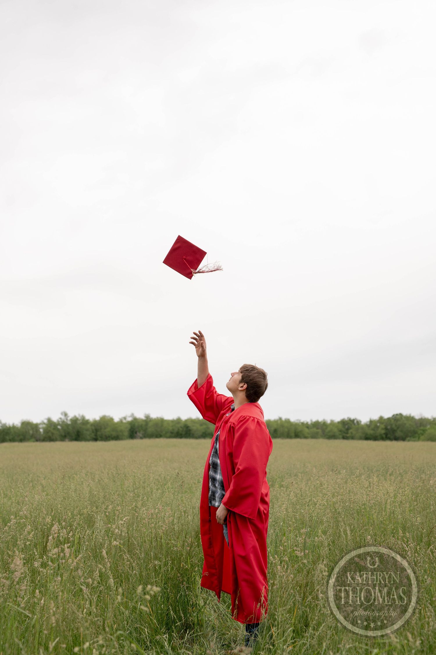 Graduate in red cap and gown throws graduation cap in an open grassy field on overcast day.
