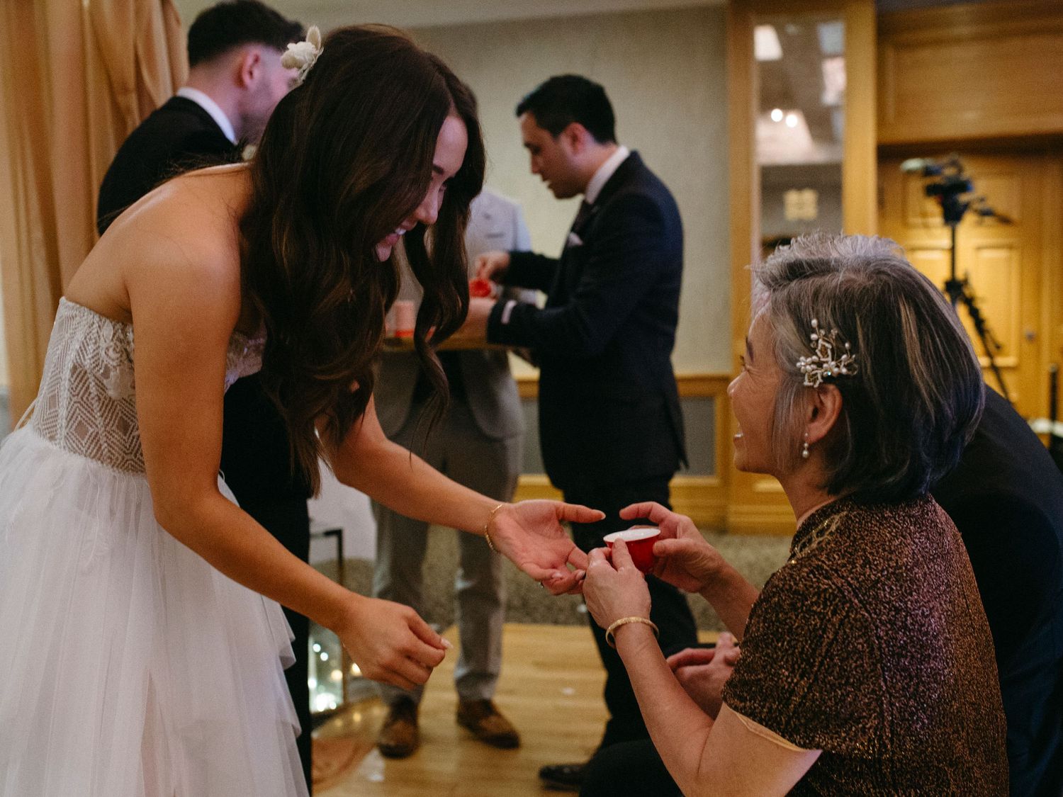 A person in a white wedding dress shares a tender moment with an elderly guest during a wedding reception.