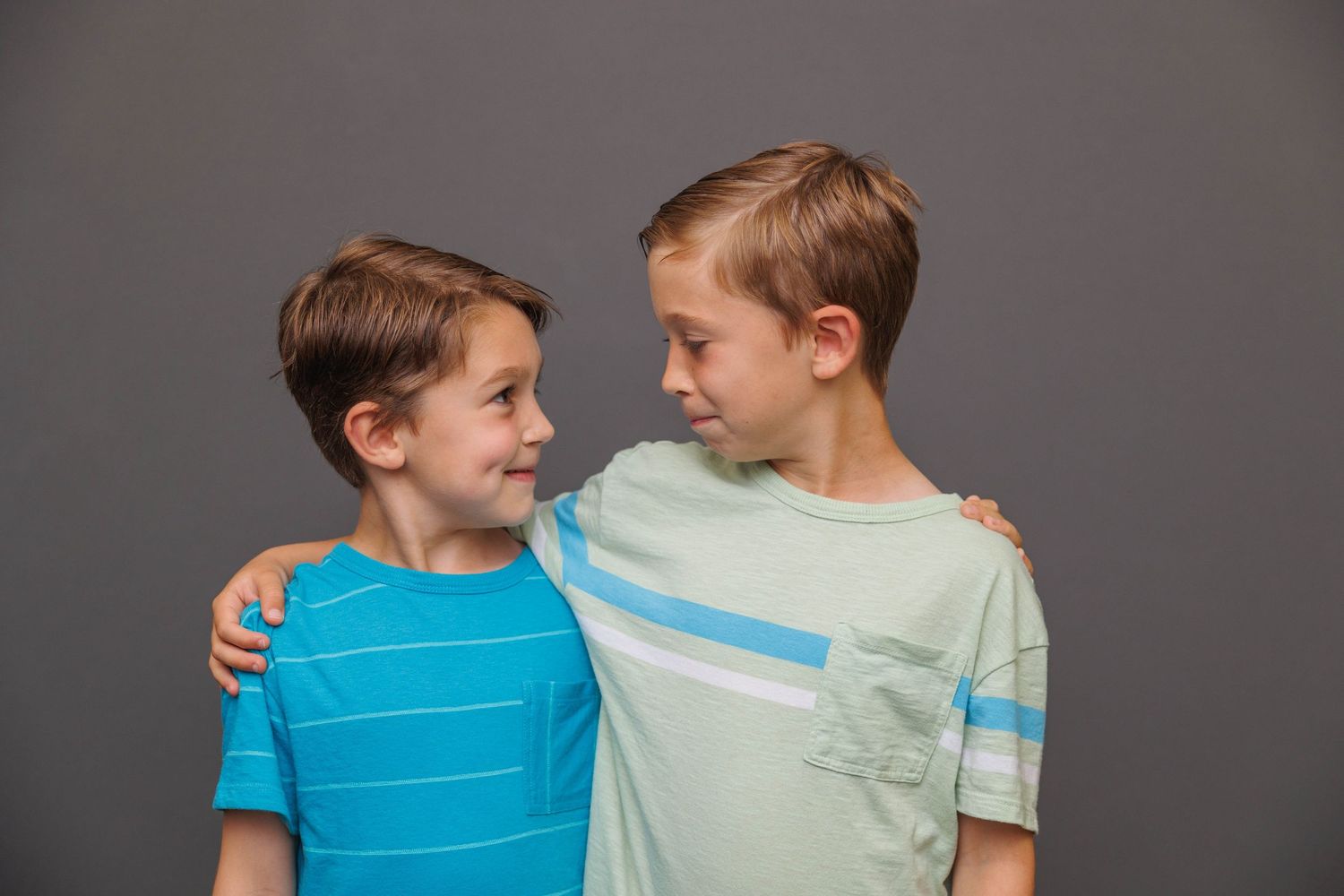 Two young friends in casual t-shirts embrace and smile at each other against a gray backdrop.