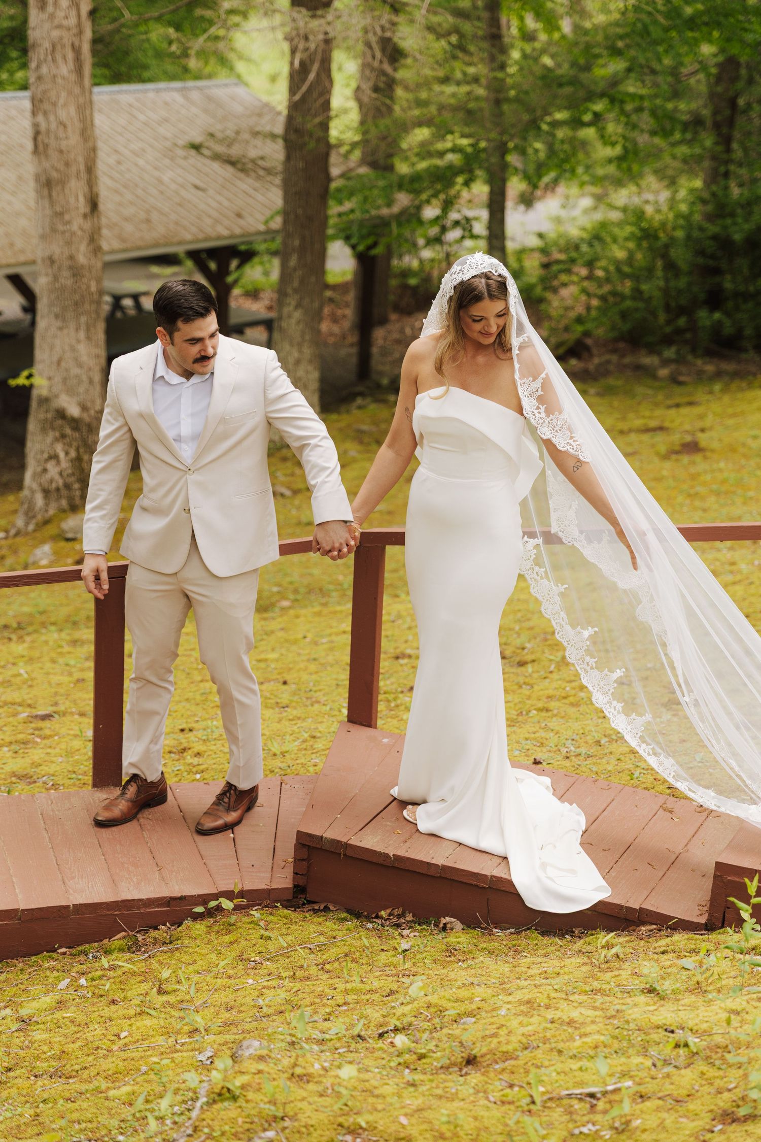 Bride in white gown with flowing veil and groom in beige suit hold hands while walking on wooden deck in outdoor forest setting.