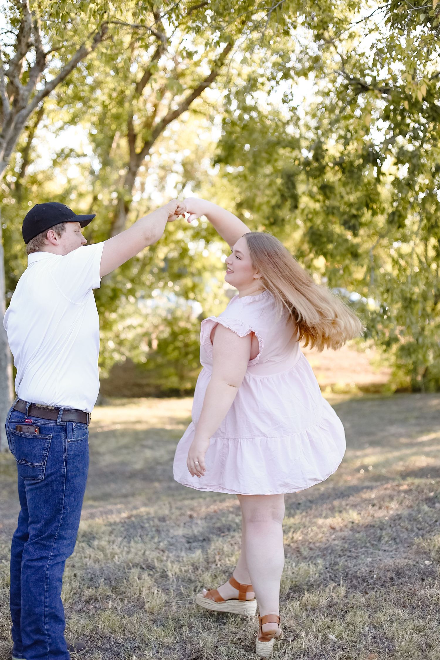 A couple dances together in a romantic twirl pose outdoors under sunlit trees in a grassy area.