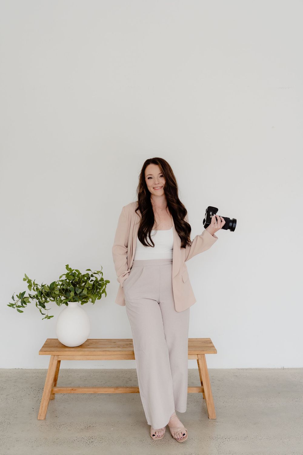 Woman in beige business attire poses against white wall with camera and decorative plant.