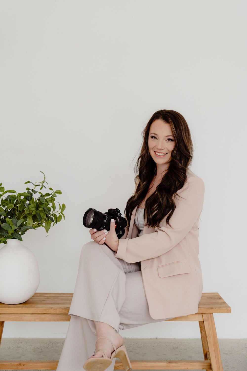 Woman in neutral toned outfit sits on wooden bench holding professional camera with potted plant nearby.