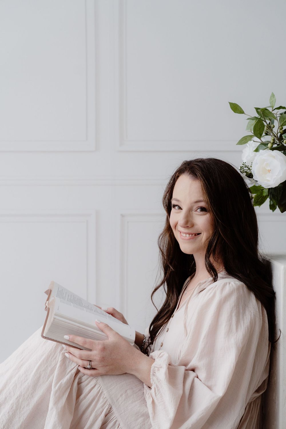 Woman in blush dress posing playfully against white wall with floral accents.