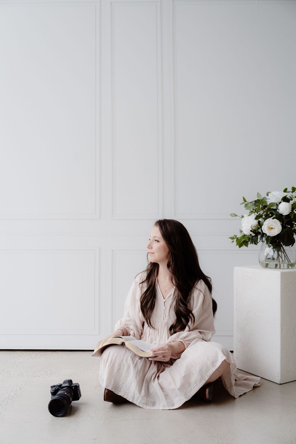 Woman in light dress sitting against white wall with minimal floral decor.