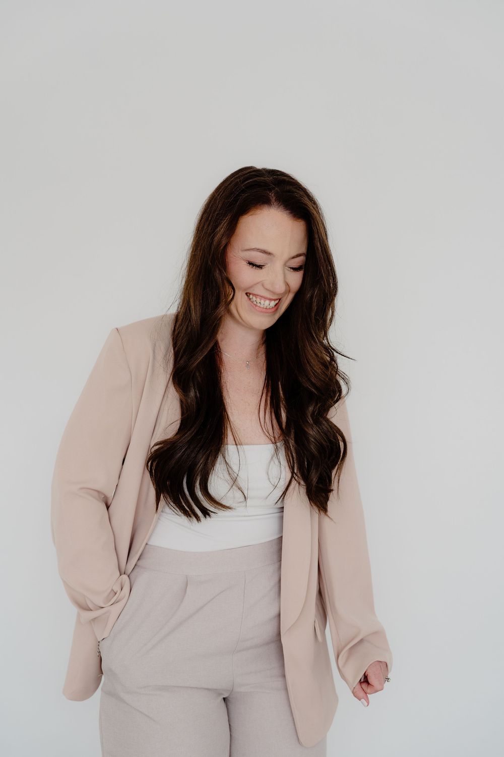 Woman in beige blazer and white top poses playfully against white background.