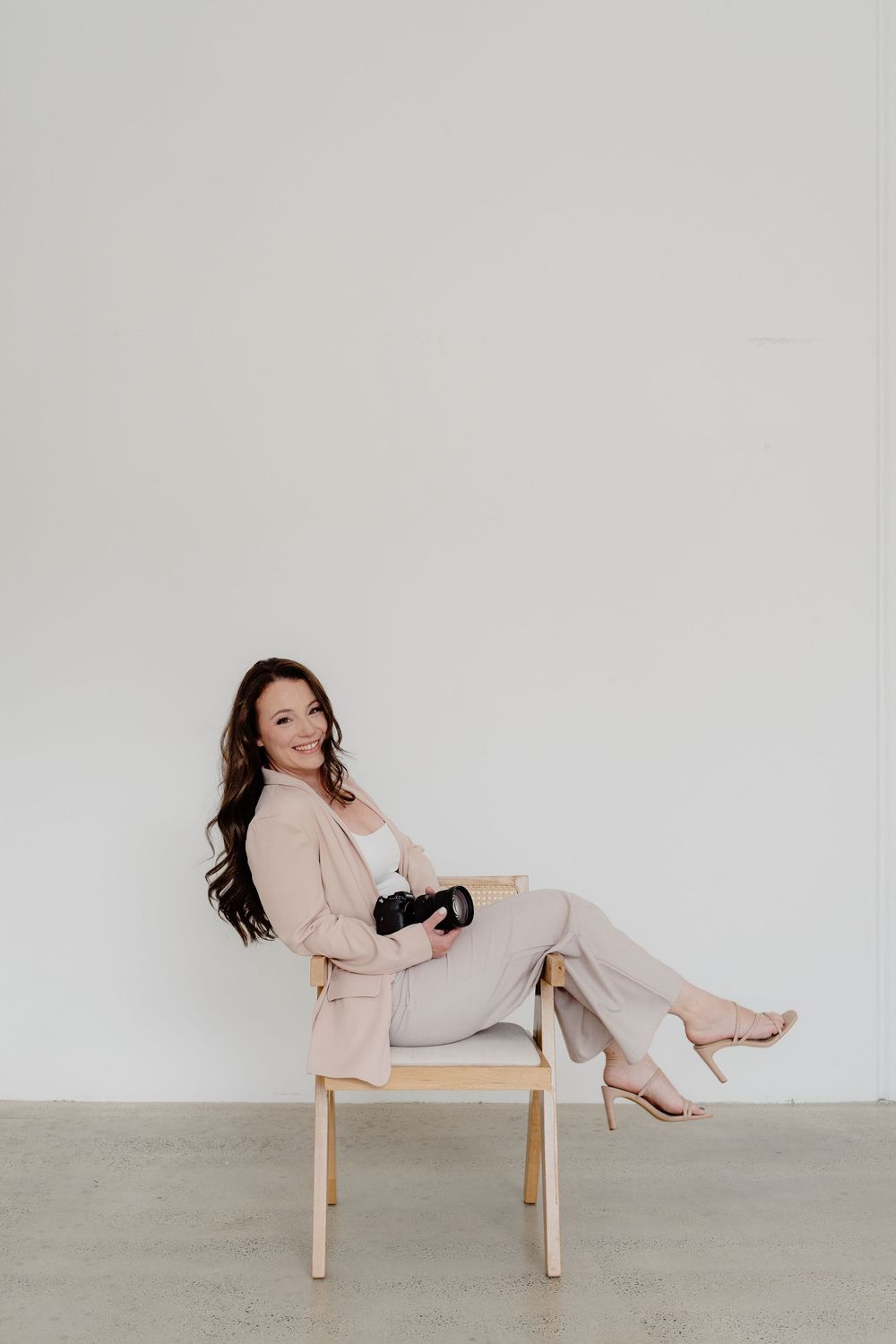 Woman in beige outfit poses elegantly on wooden chair against white backdrop.