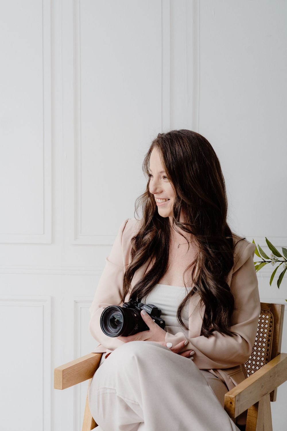 Professional photographer in beige blazer smiles while sitting in wooden chair holding camera against white wall.
