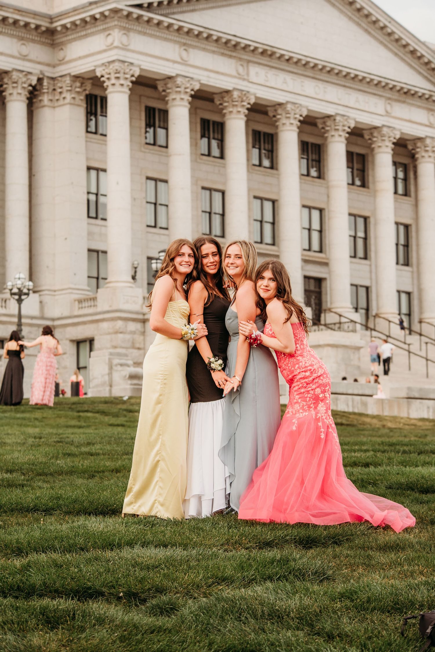 A group in formal evening gowns poses on a grassy hill in front of a grand neoclassical building with columns.