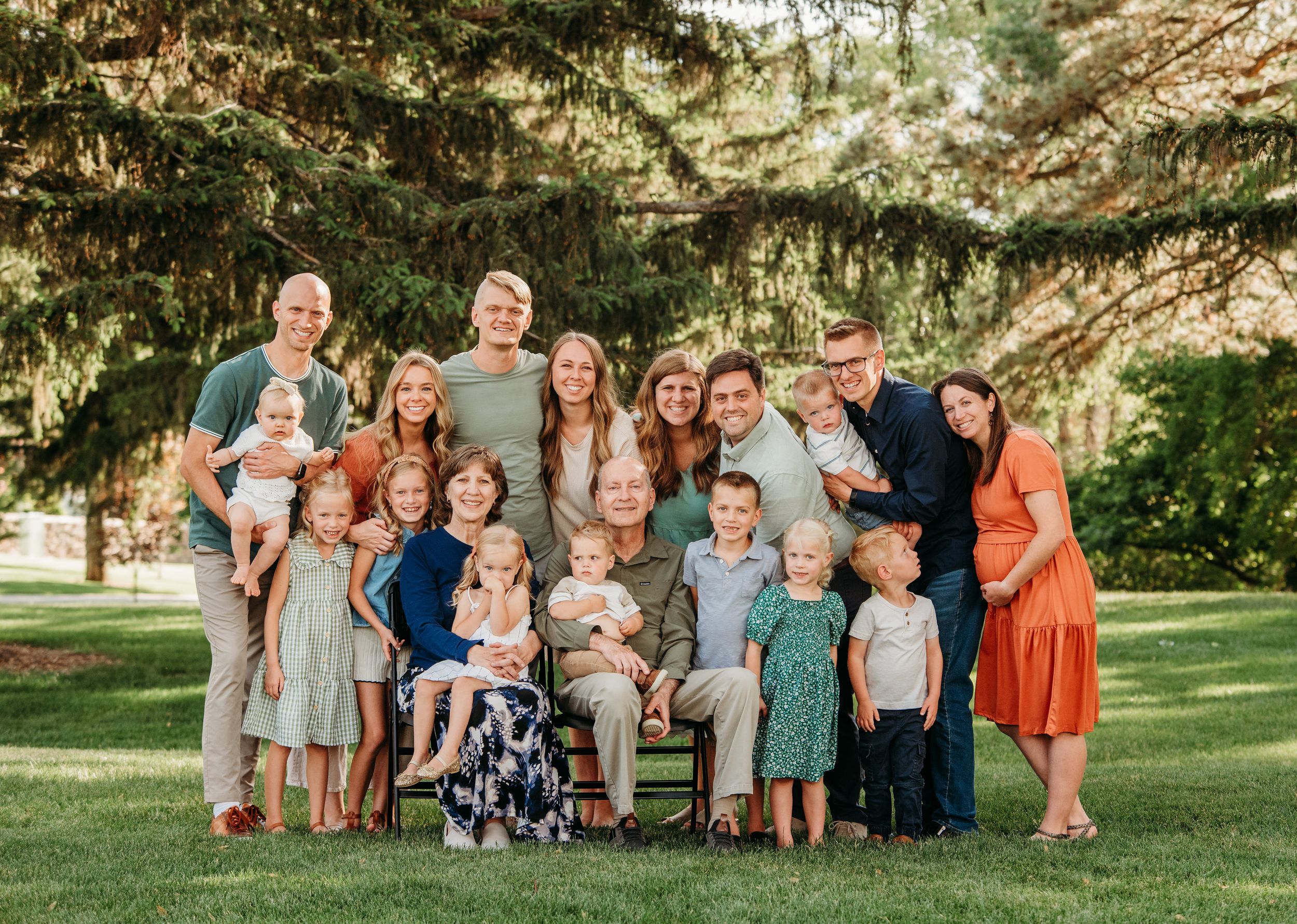 A large family group poses together outdoors on a sunny day under evergreen trees on a grassy lawn.
