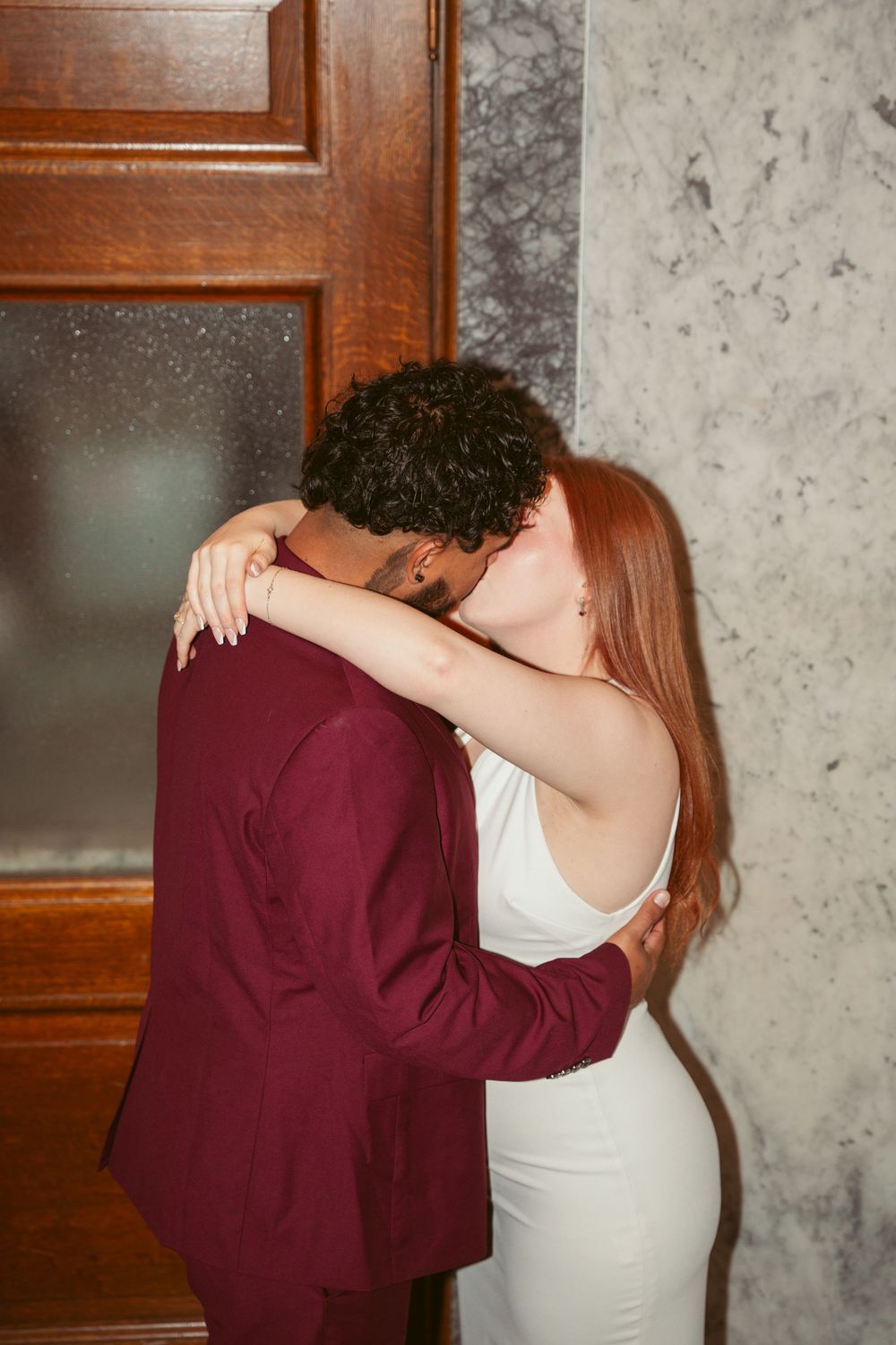 A couple shares an intimate embrace near a wooden door and marble wall, the woman wearing white and man in burgundy.