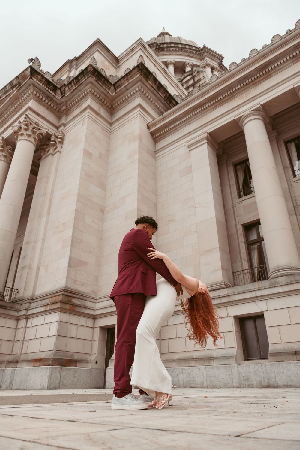 A couple in elegant attire performs a romantic dance pose in front of a grand neoclassical building with columns.